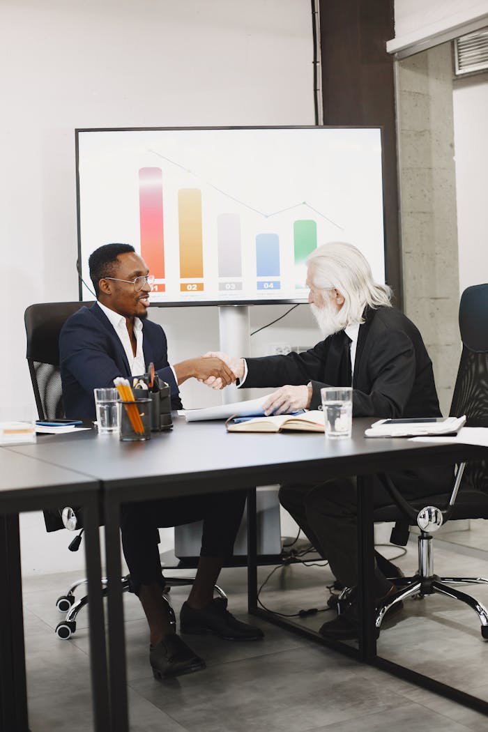Two diverse businessmen in suits shaking hands over a desk in a modern office.